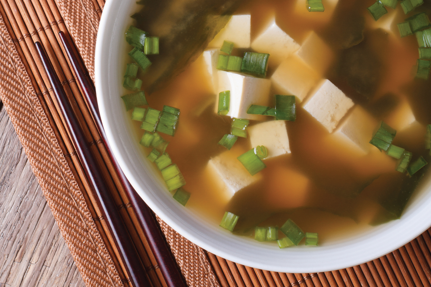 Miso Soup with Mushrooms, Bok Choy & Tofu in a white bowl placed on a white mat next to chop sticks.