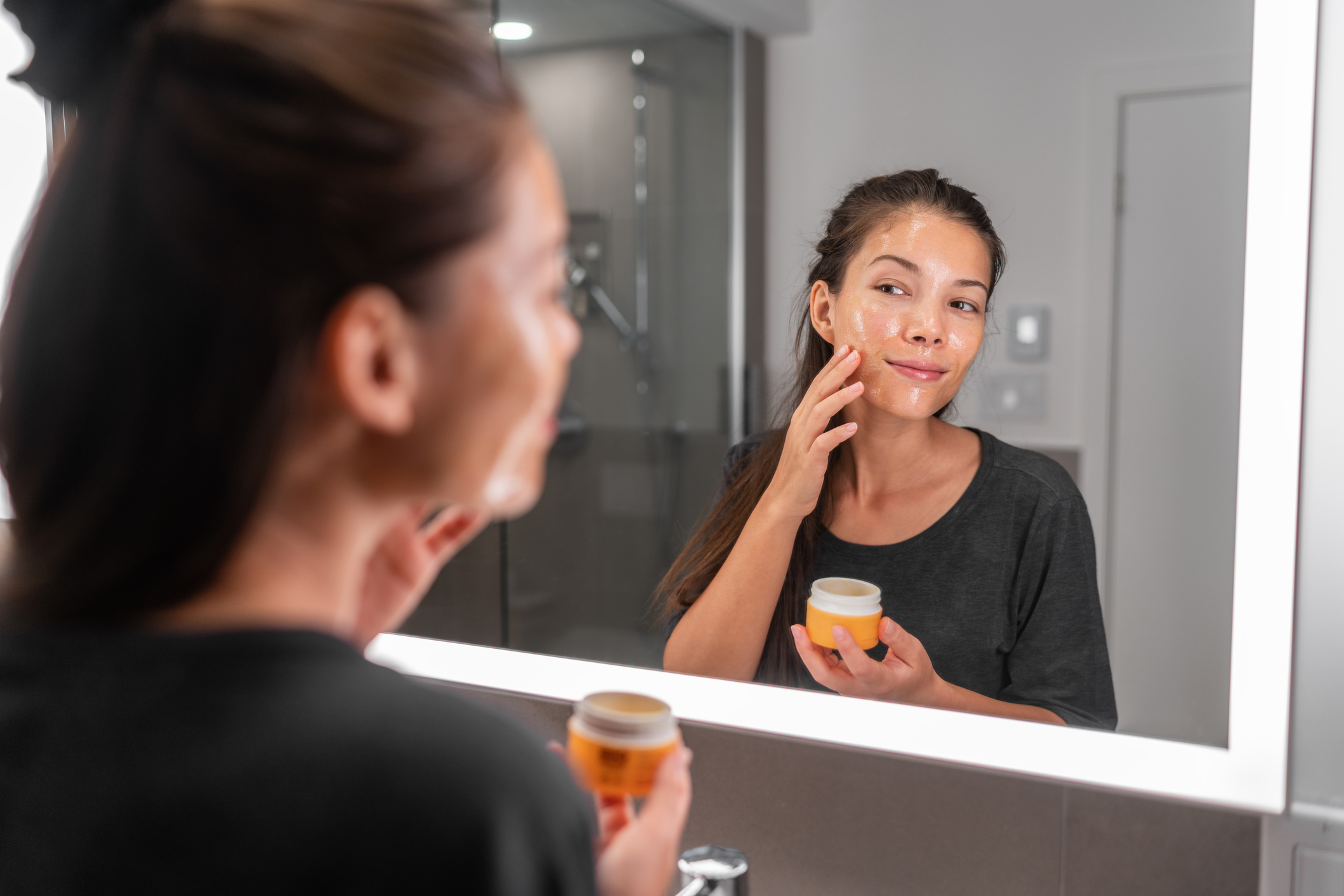 A woman adding honey to her skin in front of a bathroom mirror. 