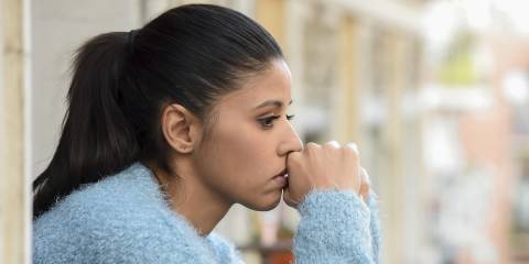 A woman sitting sadly contemplating. Hands clasped in front of mouth. 