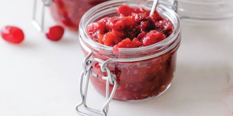 A small flip top glass jar of cranberry chutney with a small serving spoon in it, white background.