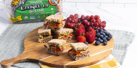 Colorful veggie sandwich on a cutting board, garnished with berries and grapes, with a package of Food for Life Ezekiel 4:9 bread in the background