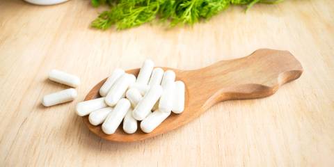 Vitamins, supplements in spoon on wooden table with green herbs in the background.